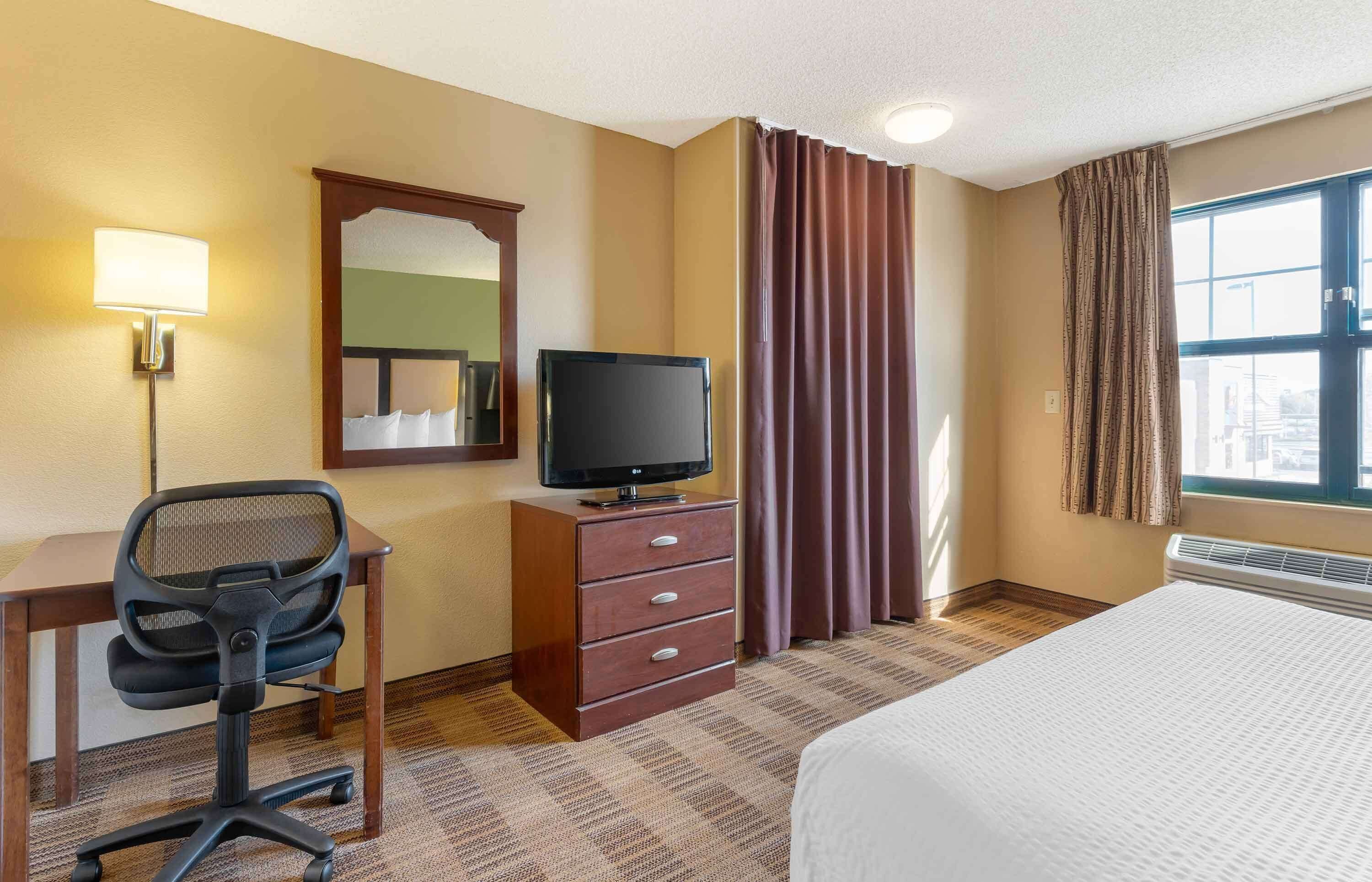 Una habitación de hotel con cama, escritorio y silla, televisor, cómoda, espejo y ventana con cortinas.