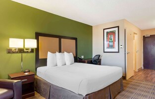 A hotel room with a large bed, two bedside lamps, a desk with a chair, a framed picture on the wall, and a door to the right.
