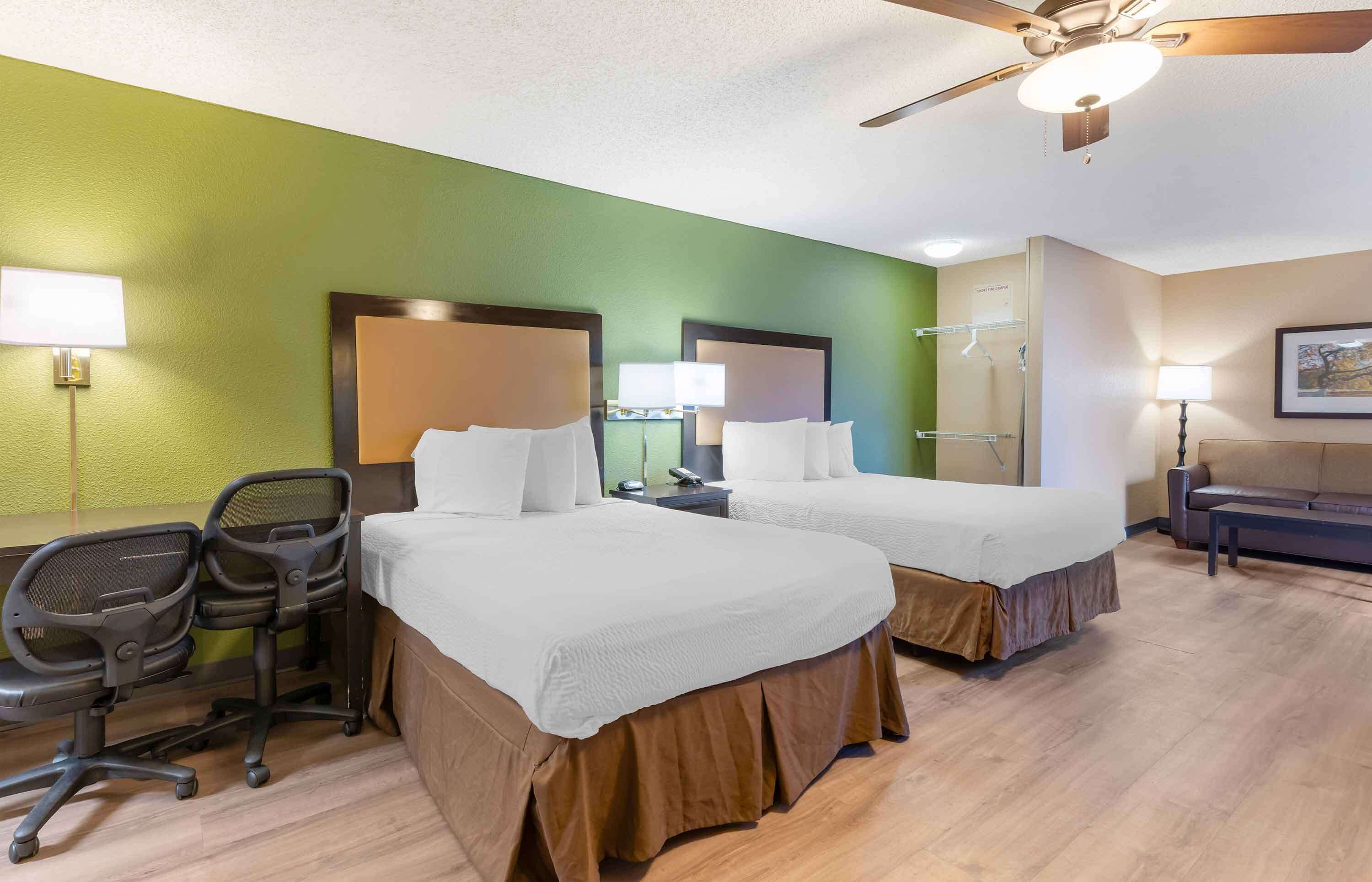 A hotel room with two beds, a desk, office chairs, a ceiling fan, and a painting on the wall.
