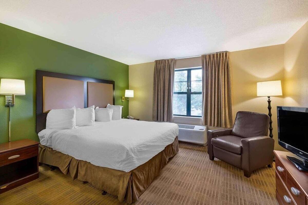 Une chambre d’hôtel avec un grand lit, une chaise, une télévision et une fenêtre avec des rideaux.
