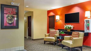 A hotel lobby with two beige armchairs, a small table with a vase of flowers, and a red wall with a framed picture and a mounted TV. - Extended Stay America Select Suites Hartford Manchester (Manchester)