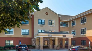 A multi-story hotel with a beige facade, a lobby entrance, and a parking area with two cars. - Extended Stay America Select Suites Hartford Manchester (Manchester)