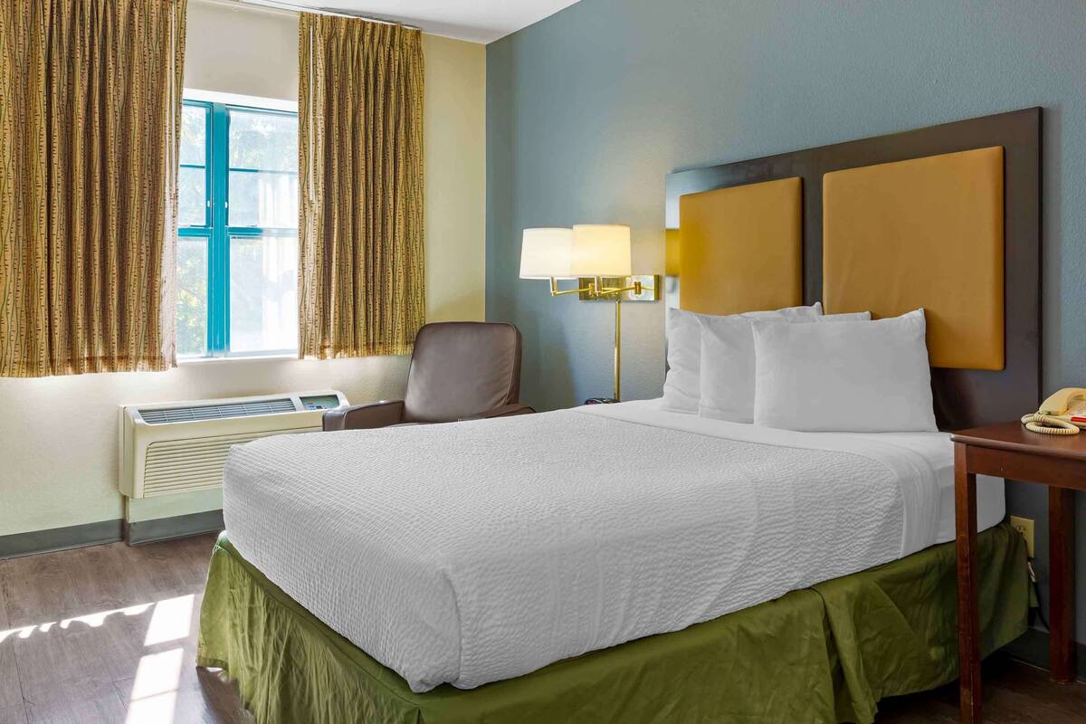 A hotel room with a bed, a chair, a desk with a lamp, and a window with curtains.