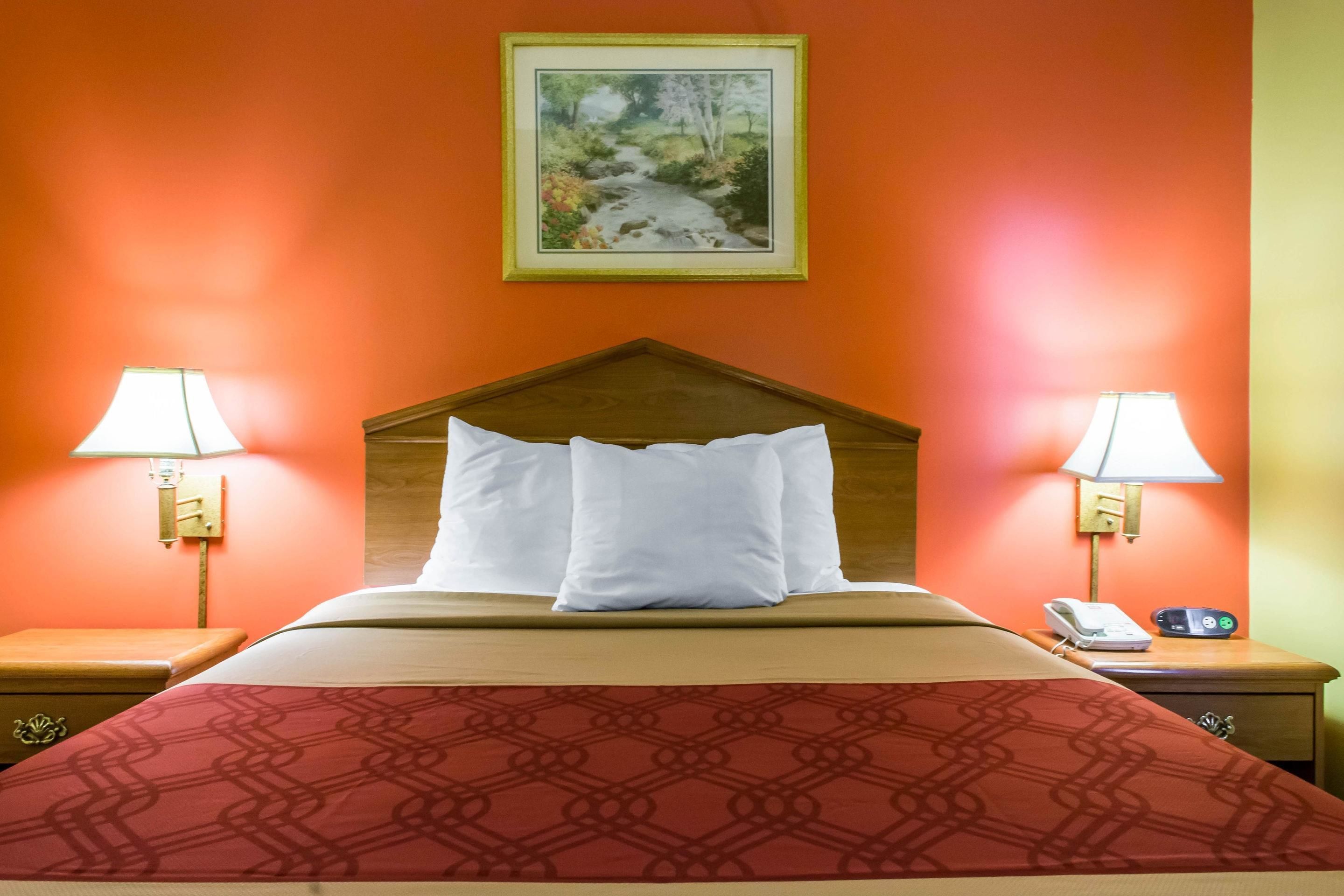 A hotel room with a bed, two bedside lamps, a nightstand with a phone and a clock, and a framed picture on the wall.