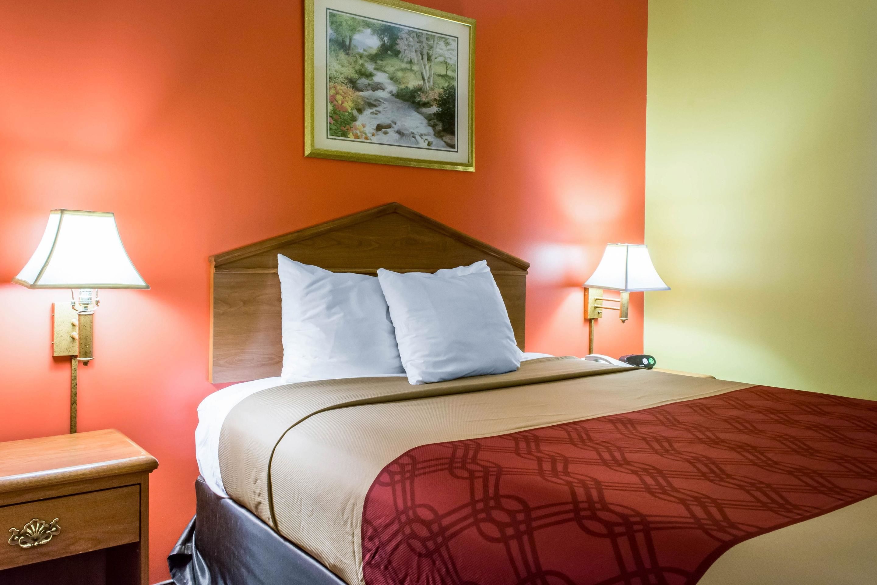 A hotel room with a bed, two bedside lamps, a wooden headboard, and a framed picture on the wall.