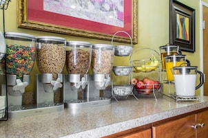 Una barra de desayuno con dispensadores de cereales, una cesta de fruta y una jarra de zumo.
