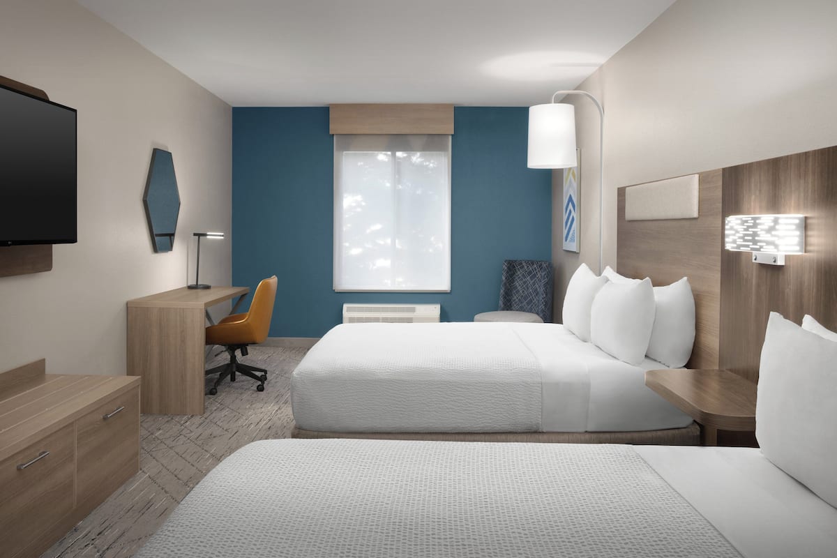 Une chambre d’hôtel avec deux lits, un bureau, une chaise, une télévision et une fenêtre avec des stores.