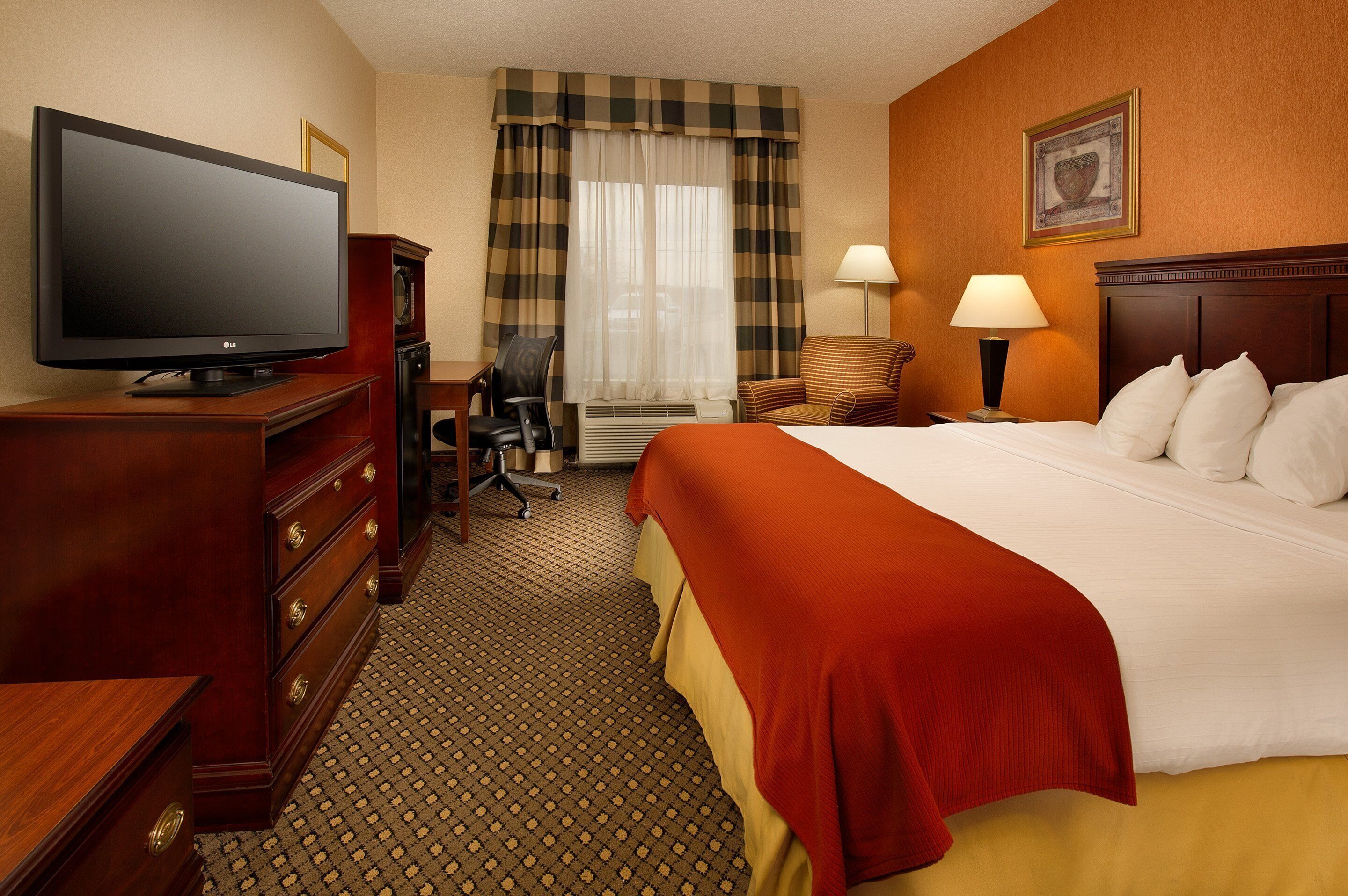 A hotel room with a bed, a television, a desk, a chair, a lamp, and a window with curtains.