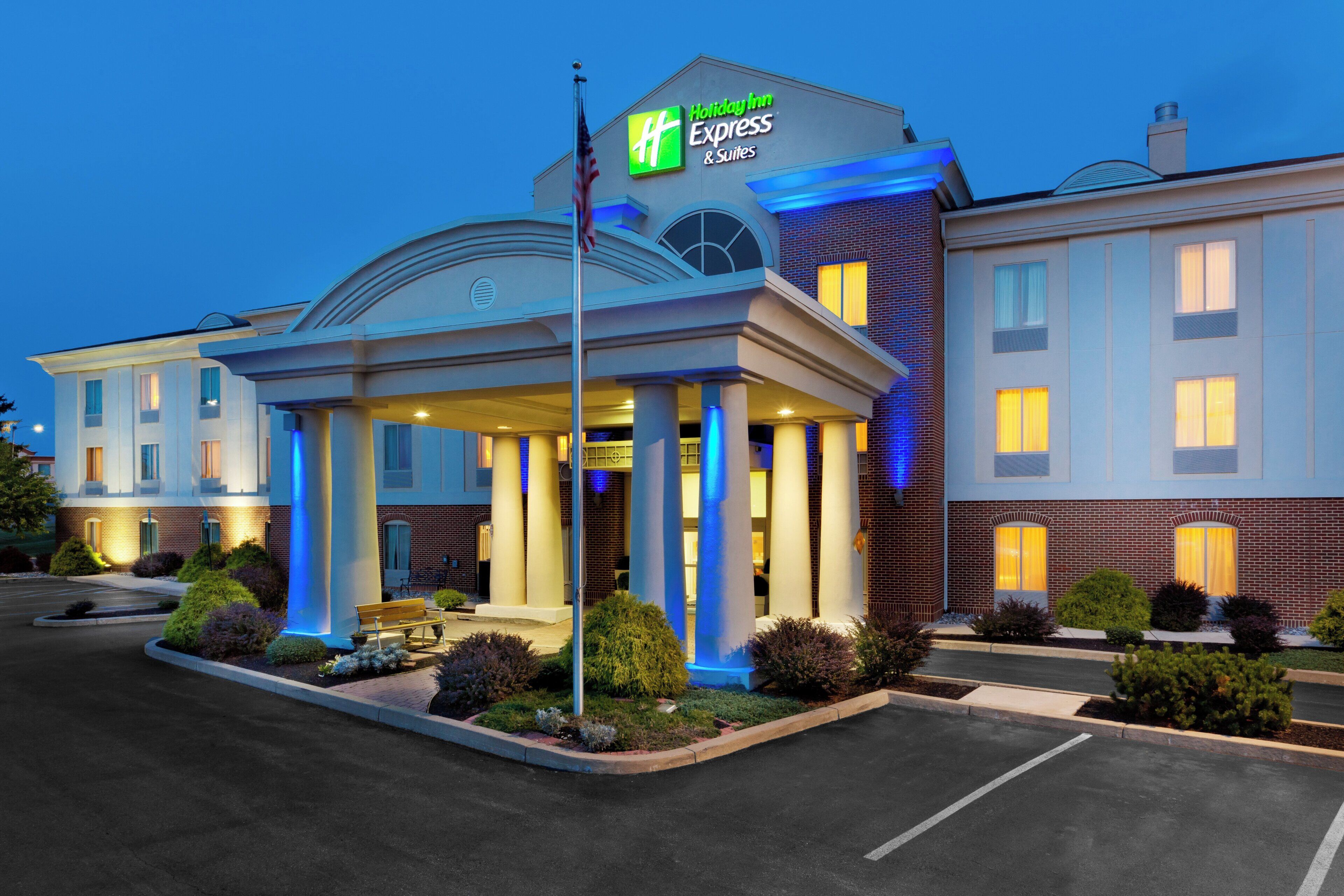 A Holiday Inn Express & Suites hotel with a covered entrance, outdoor seating, and a flagpole.