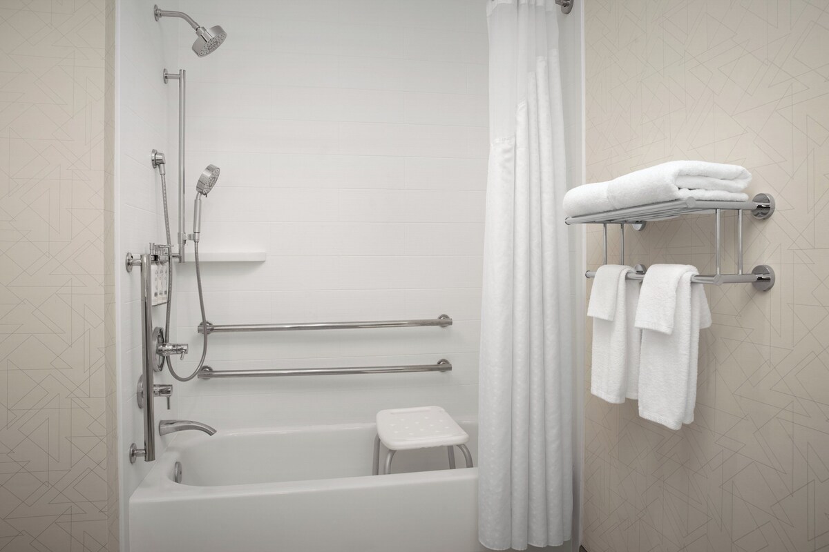 A modern bathroom with a bathtub, shower, grab bars, a small stool, and a towel rack.