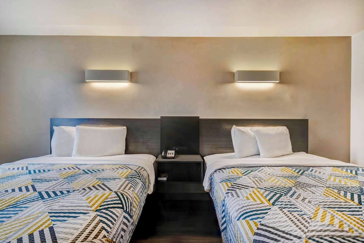 A hotel room with two beds, each with a patterned bedspread, a nightstand with a phone, and wall-mounted lighting.