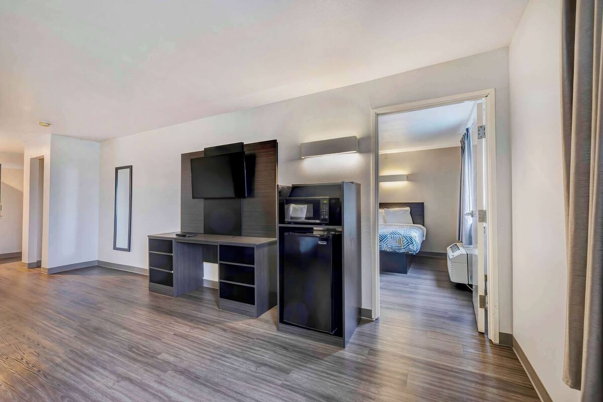 A modern hotel room with a flat-screen TV, a mini-fridge, and a microwave. The room has a wooden floor and a bed with a blue and white patterned bedspread.