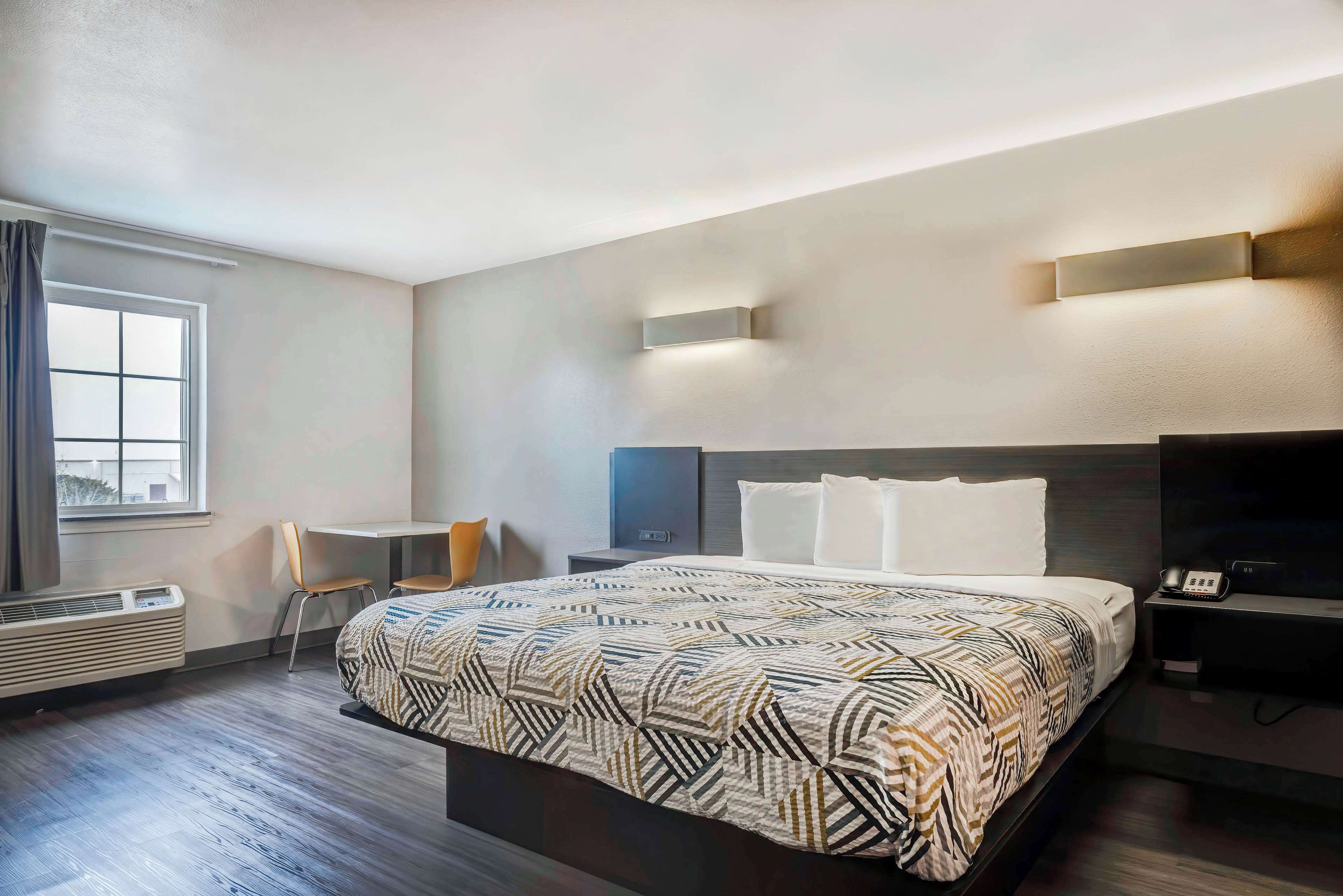 A modern hotel room with a large bed, a desk, and a chair. There is a window with curtains, a heater, and a wall-mounted phone.