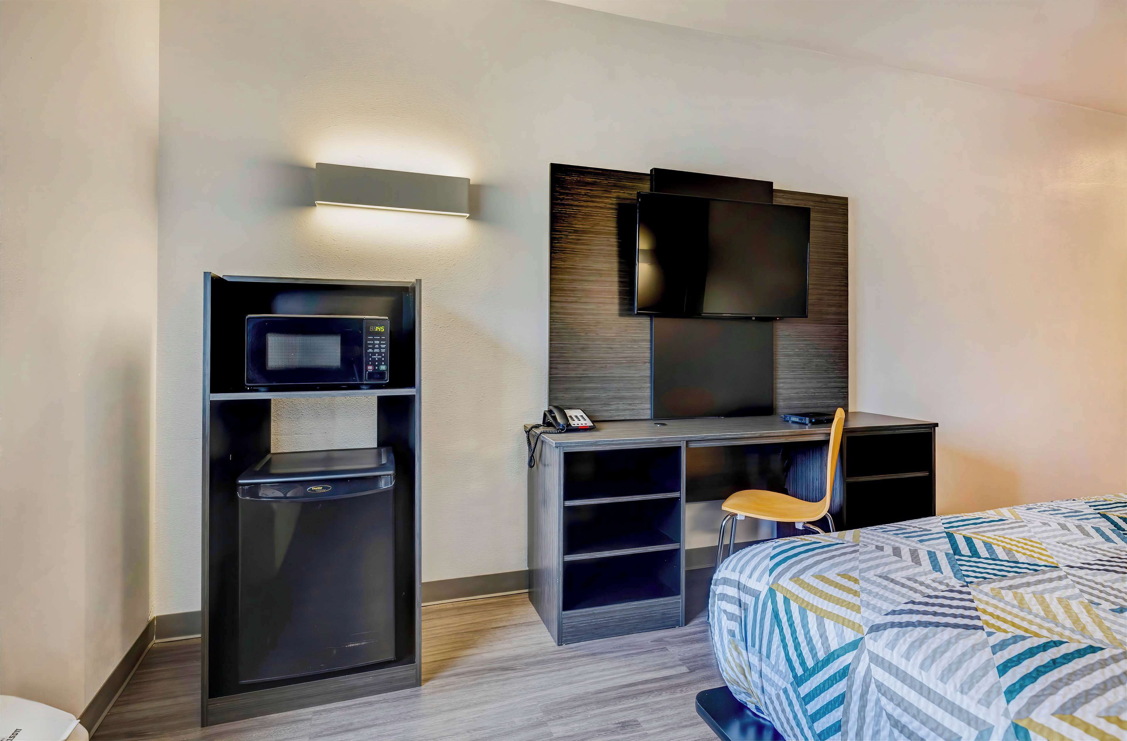 A hotel room with a kitchenette, a flat-screen TV mounted on the wall, a bed with a patterned bedspread, and a desk with a chair.