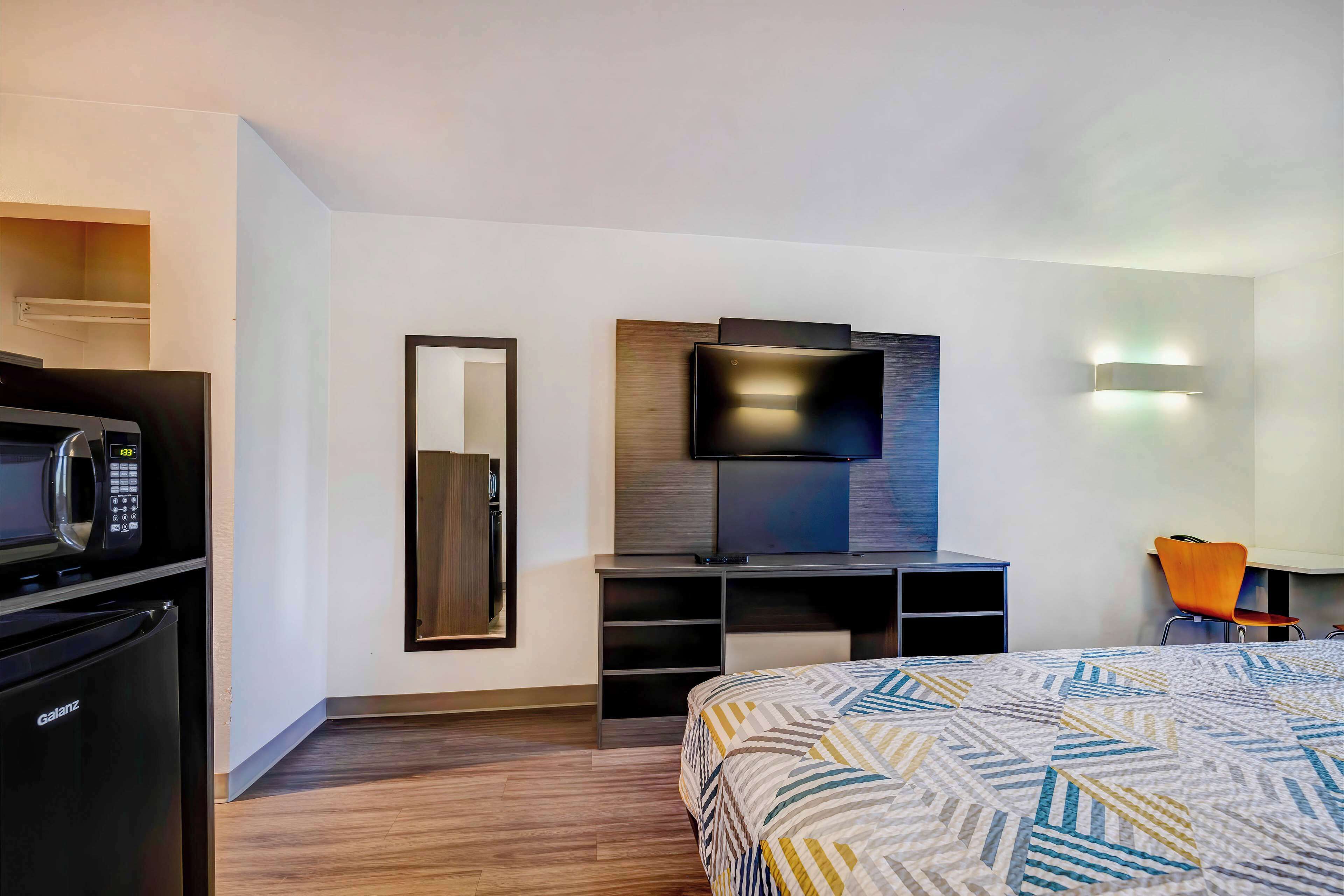 Une chambre d’hôtel moderne équipée d’un lit, d’un téléviseur à écran plat fixé au mur, d’un four à micro-ondes, d’un réfrigérateur et d’un bureau avec une chaise.