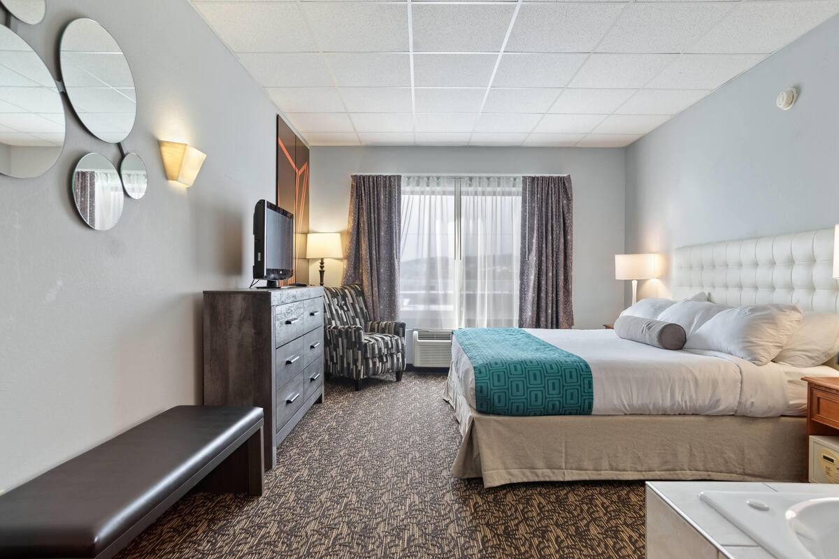 Ein Hotelzimmer mit Bett, Sitzbank, Kommode, Fernseher, einem Fenster mit Vorhängen und einem wandmontierten Spiegel.