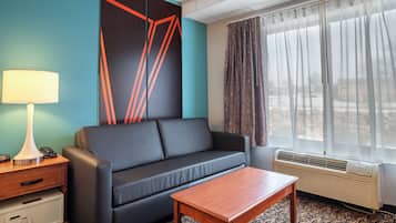 A modern hotel room with a dark sofa, a wooden coffee table, a lamp, a telephone, a safe, and a large window with curtains.