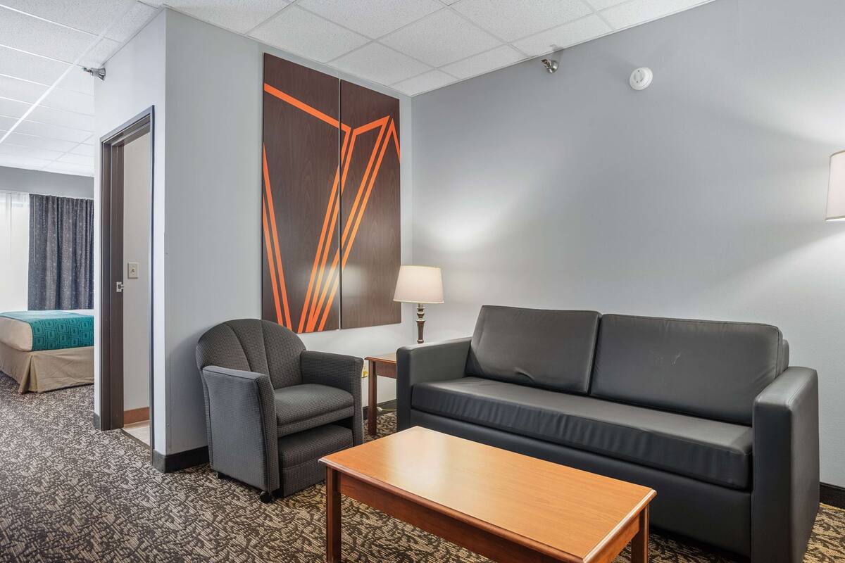 Ein Hotelzimmer mit einer Couch, einem Sessel und einem kleinen Tisch. Eine Tür führt in einen weiteren Raum, und ein Fenster mit Vorhängen ist vorhanden.
