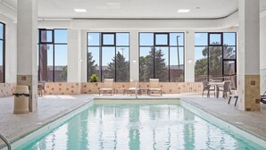 An indoor swimming pool with clear views through large windows, a tiled floor, and a seating area with chairs and tables. - Elevation Inn & Suites, Trademark Collection by Wyndham (Rapid City)
