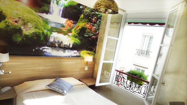 Una habitación de hotel con una cama, una mesita de noche, una lámpara, un mural grande en la pared, una ventana con balcón y una puerta.