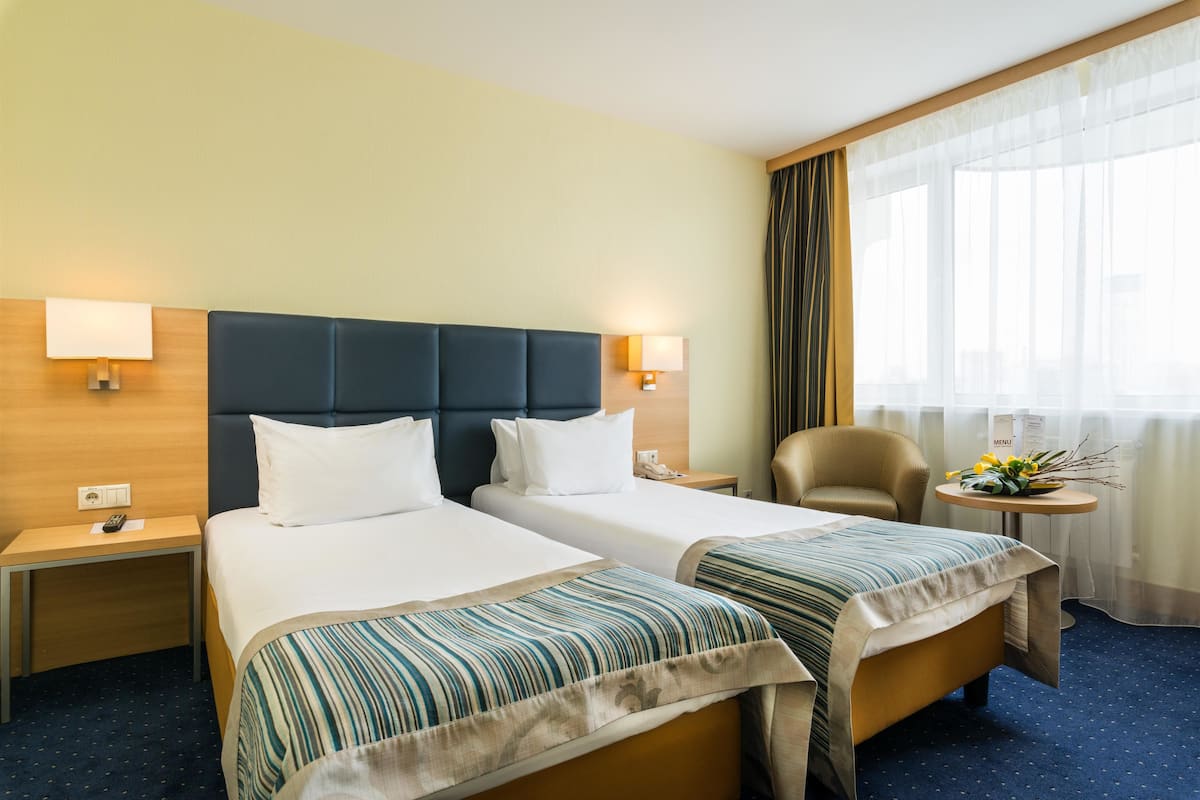 A hotel room with two beds, a small table with a lamp, a chair, and a window with curtains.