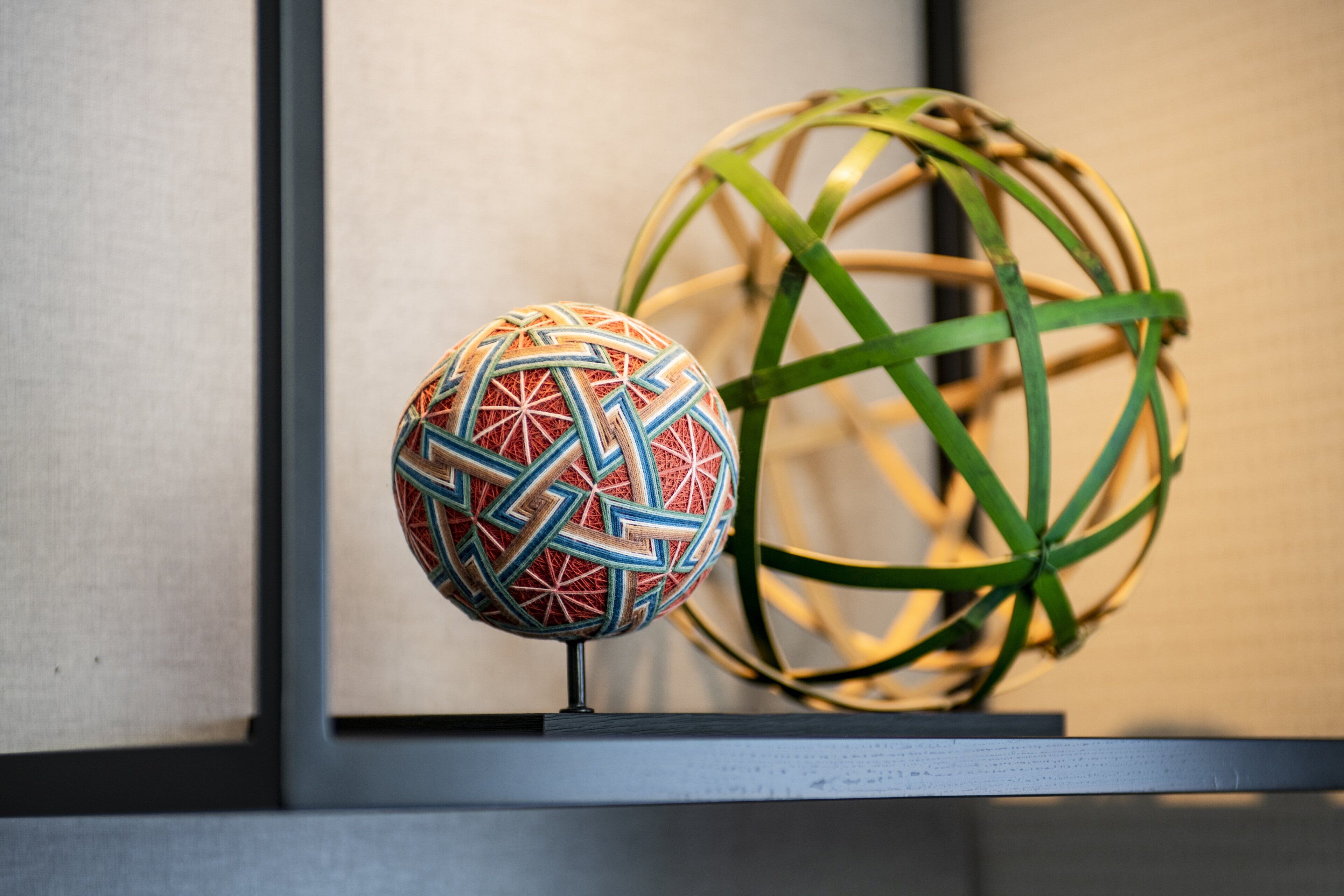 Dos objetos esféricos con patrones geométricos y un objeto esférico verde con diseño tejido, exhibidos sobre una estantería frente a una pared.