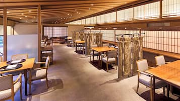A spacious dining area with multiple tables and chairs, wooden beams on the ceiling, and large windows with a view of the outdoors.