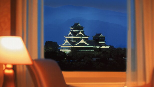 A view of a traditional Japanese castle at night through a window, with a lamp and chair in the foreground. - Hotel Nikko Kumamoto (Kumamoto)