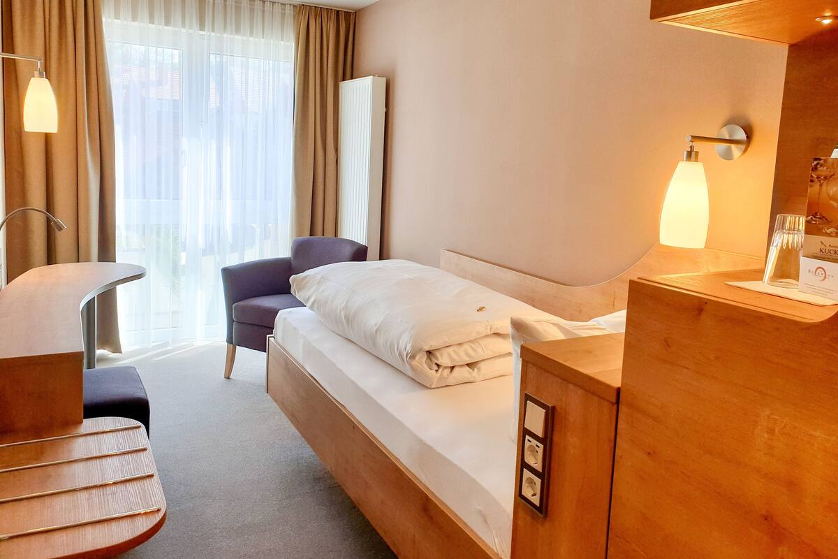 A hotel room with a bed, a chair, a desk, a lamp, and a window with curtains.