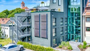 A modern building with a glass facade, a metal staircase, and a red-roofed tower in the background. - Hotel Beckmann (Goettingen)