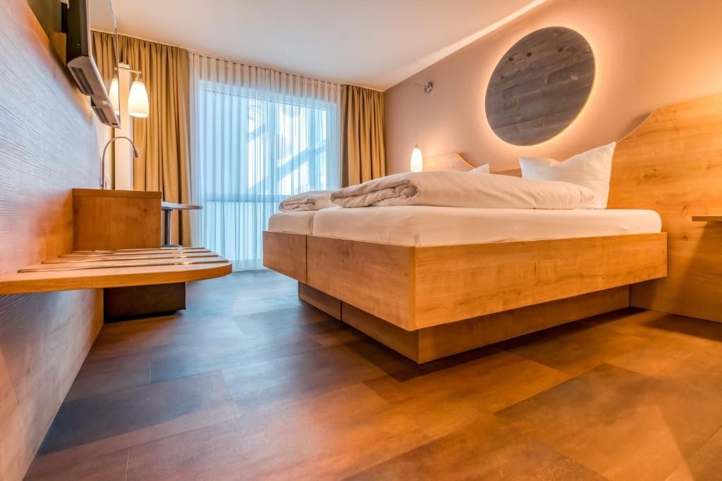 A modern hotel room with a bed, a wooden bedside table, a built-in bench, and a large window with curtains.