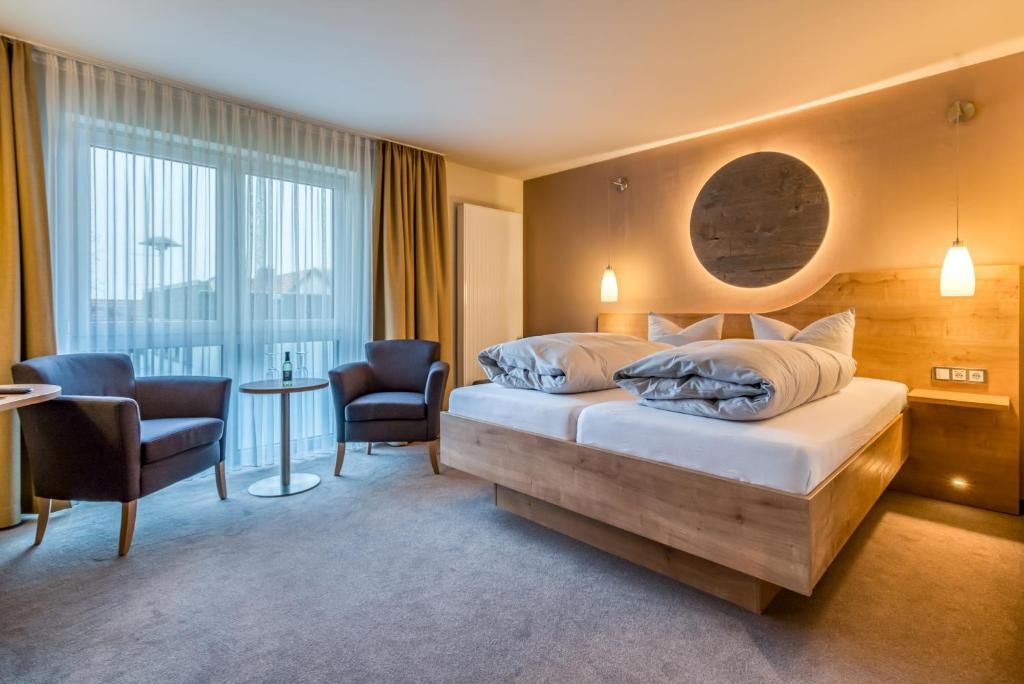 A hotel room with a bed, two armchairs, a small table, and a large window with curtains.