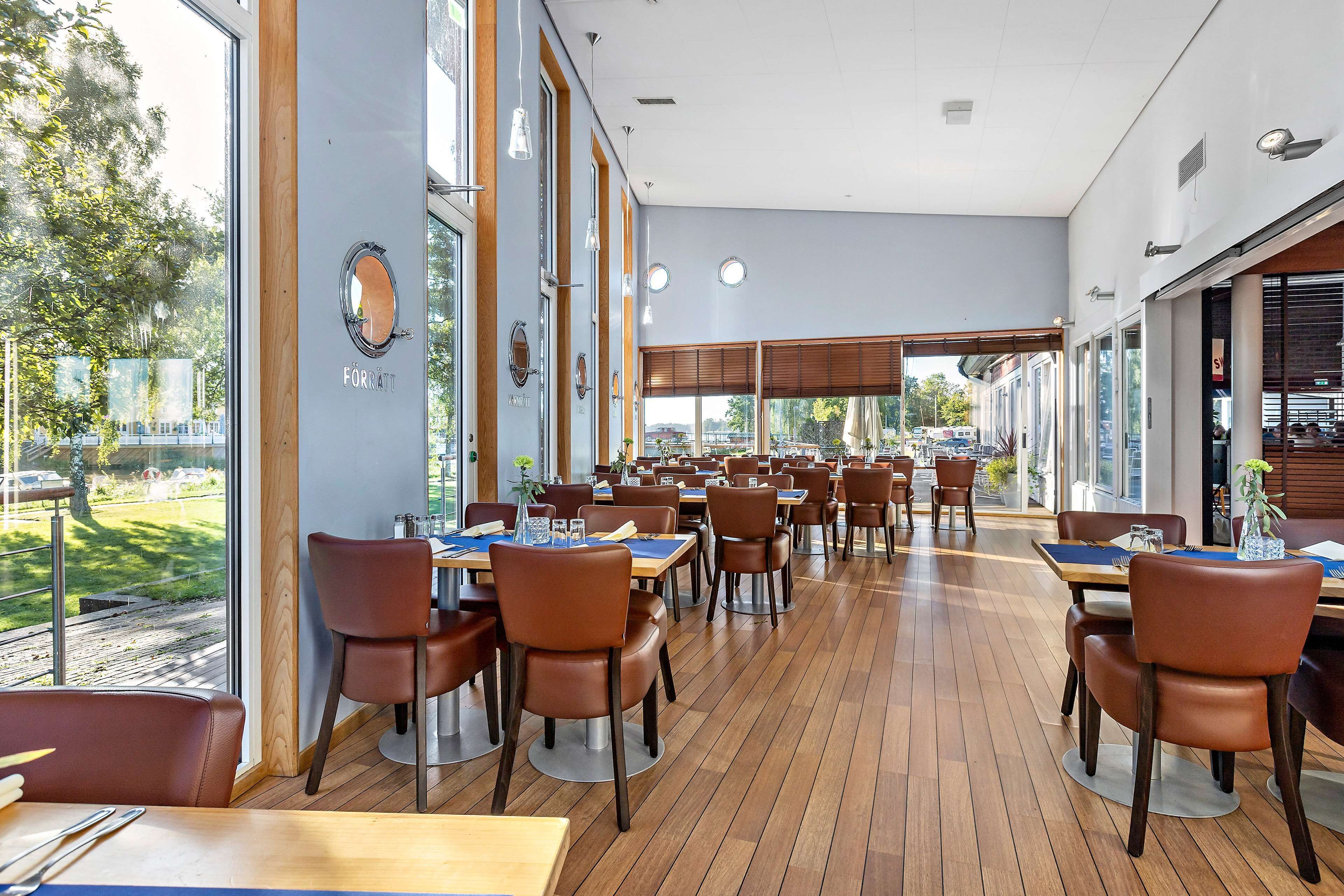 A spacious indoor dining area with wooden flooring and brown leather chairs. Tables are set with blue placemats and white tableware. Large windows provide views of the outdoors.