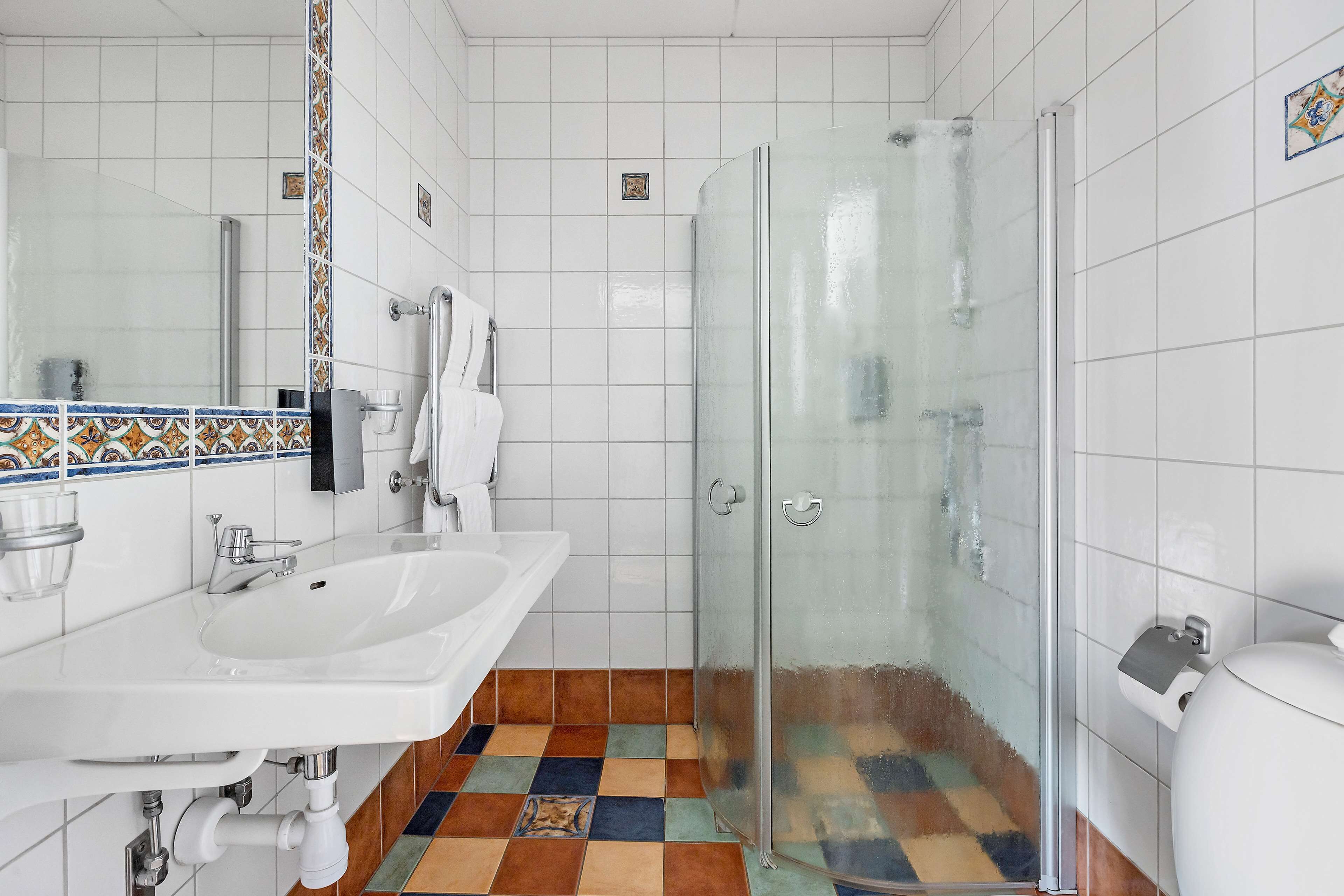 Een badkamer met een wit wastafelmeubel, een douchecabine van glas en een betegelde vloer met een kleurrijk patroon.