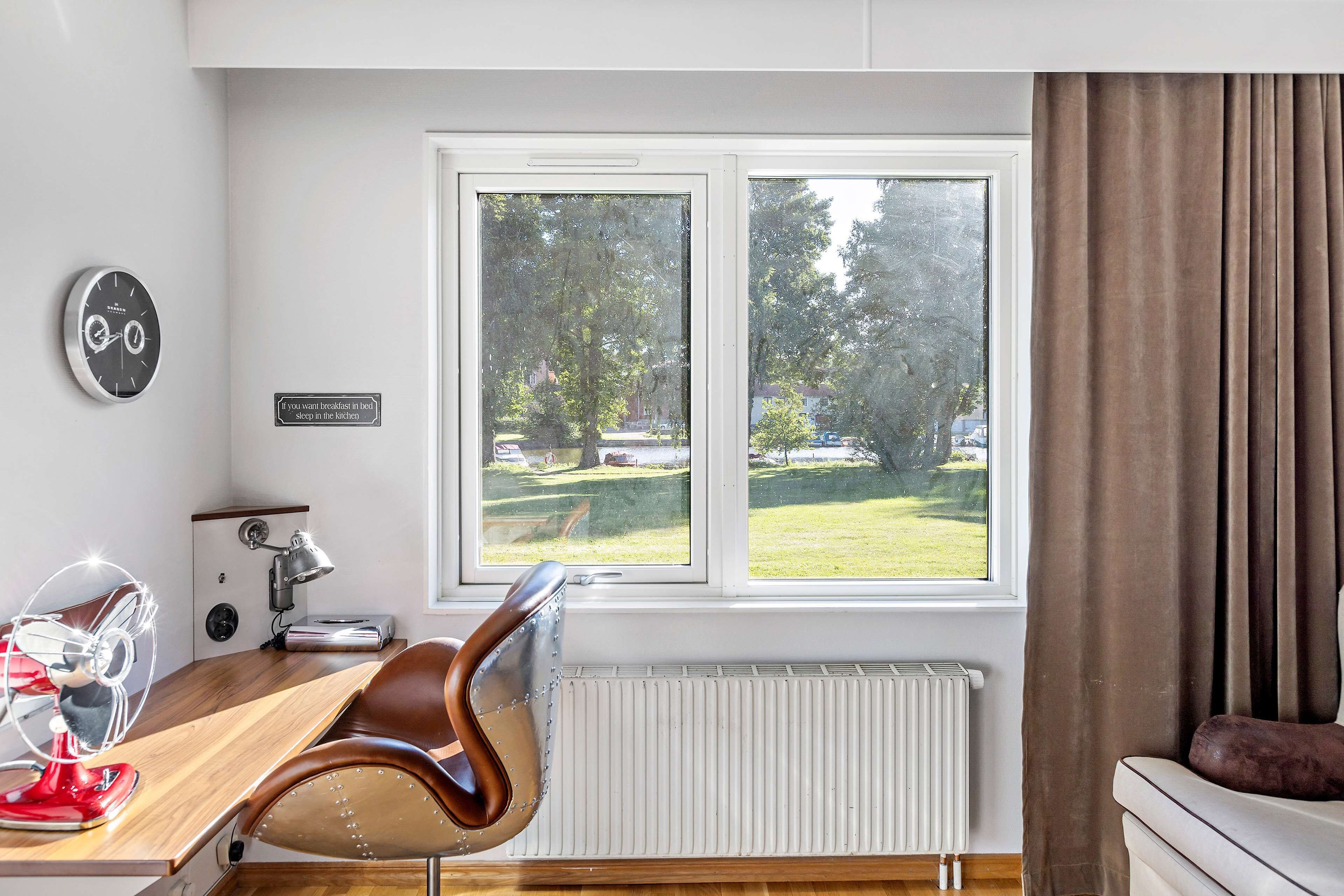 A modern room with a wooden desk, a red fan, a clock, a window with a view of a green landscape, and a white radiator.
