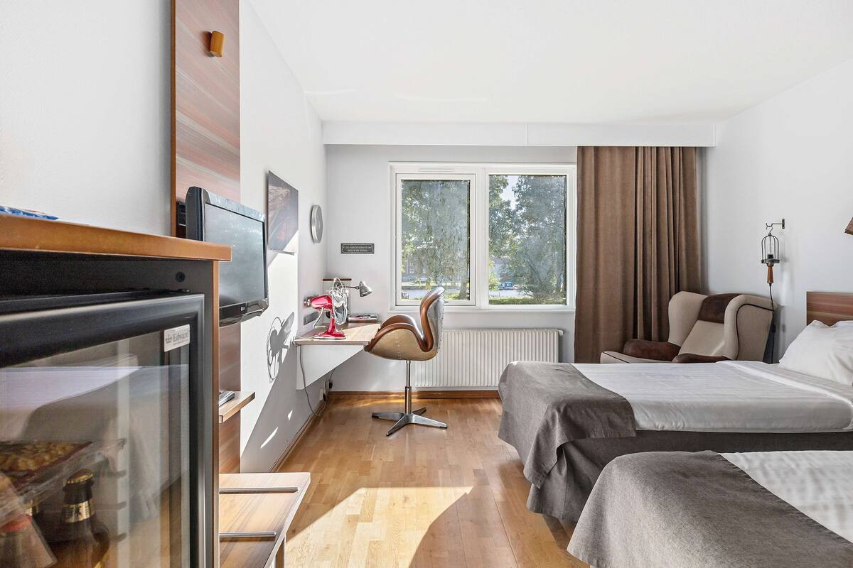 Una habitación de hotel con cama, escritorio con silla, refrigerador y ventana con cortinas.