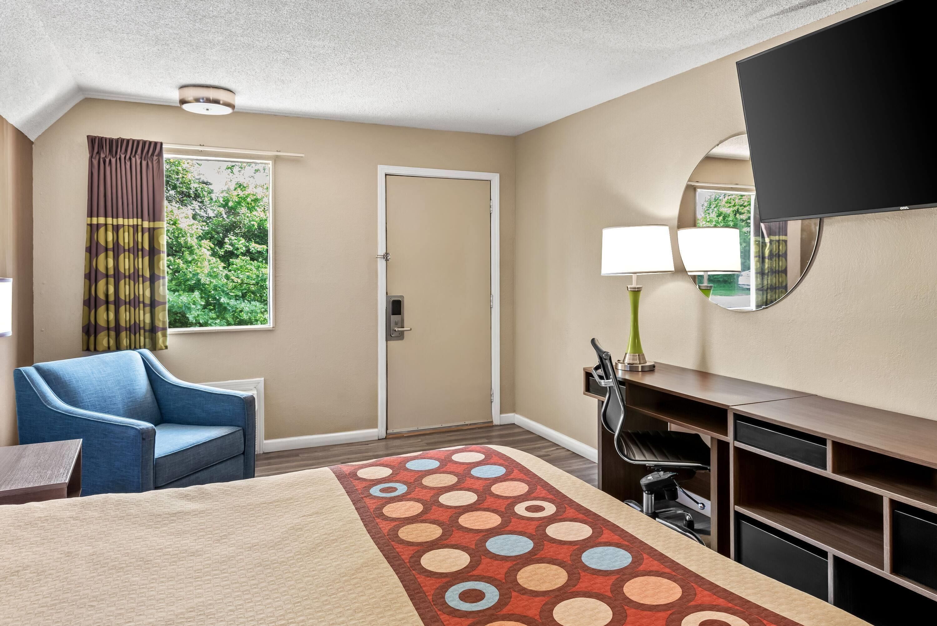 Une chambre d’hôtel avec un lit, un fauteuil bleu, un bureau avec une lampe, une télévision et une fenêtre avec des rideaux.
