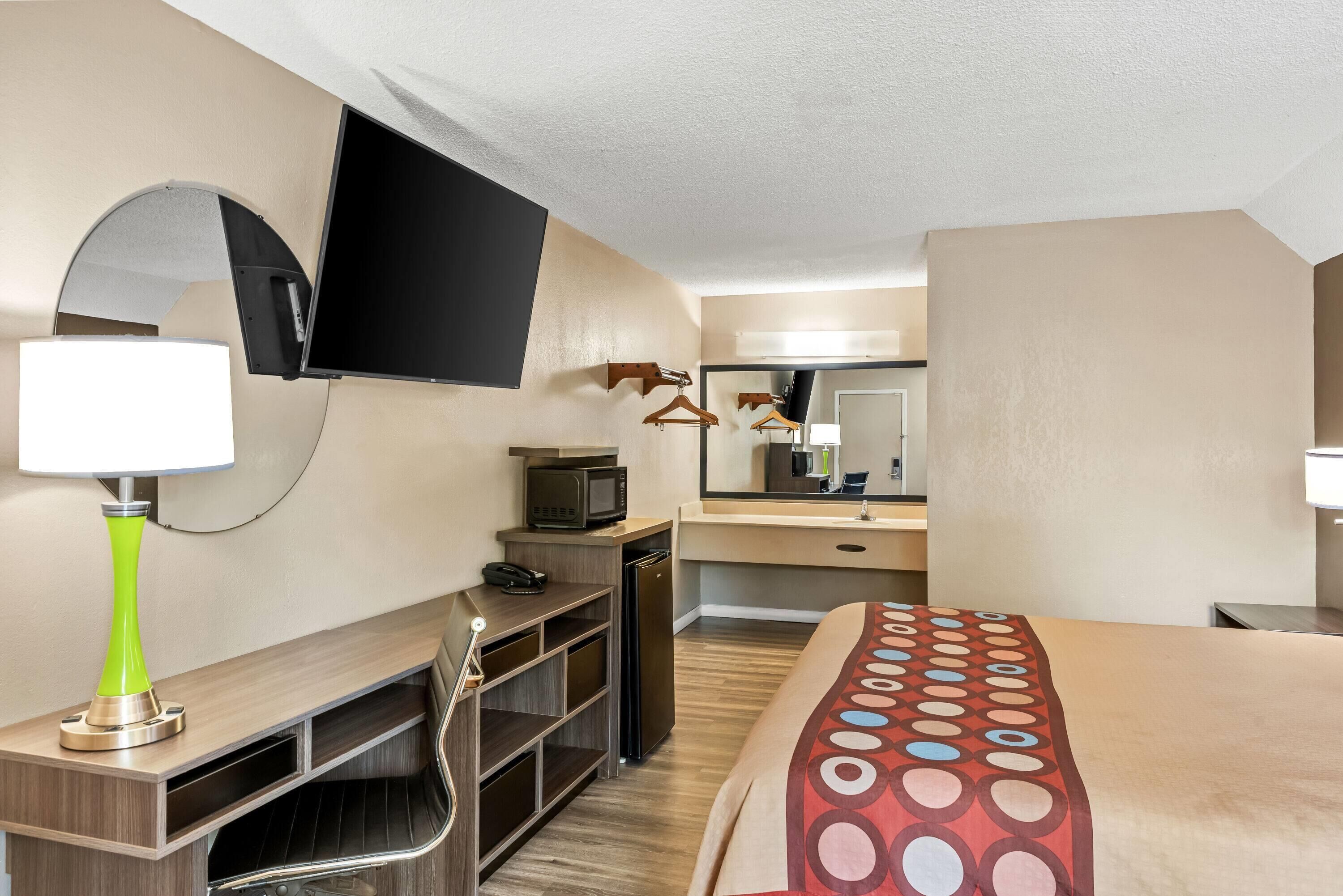 Una habitación de hotel con cama, escritorio con lámpara, televisor, microondas, refrigerador pequeño y espejo.