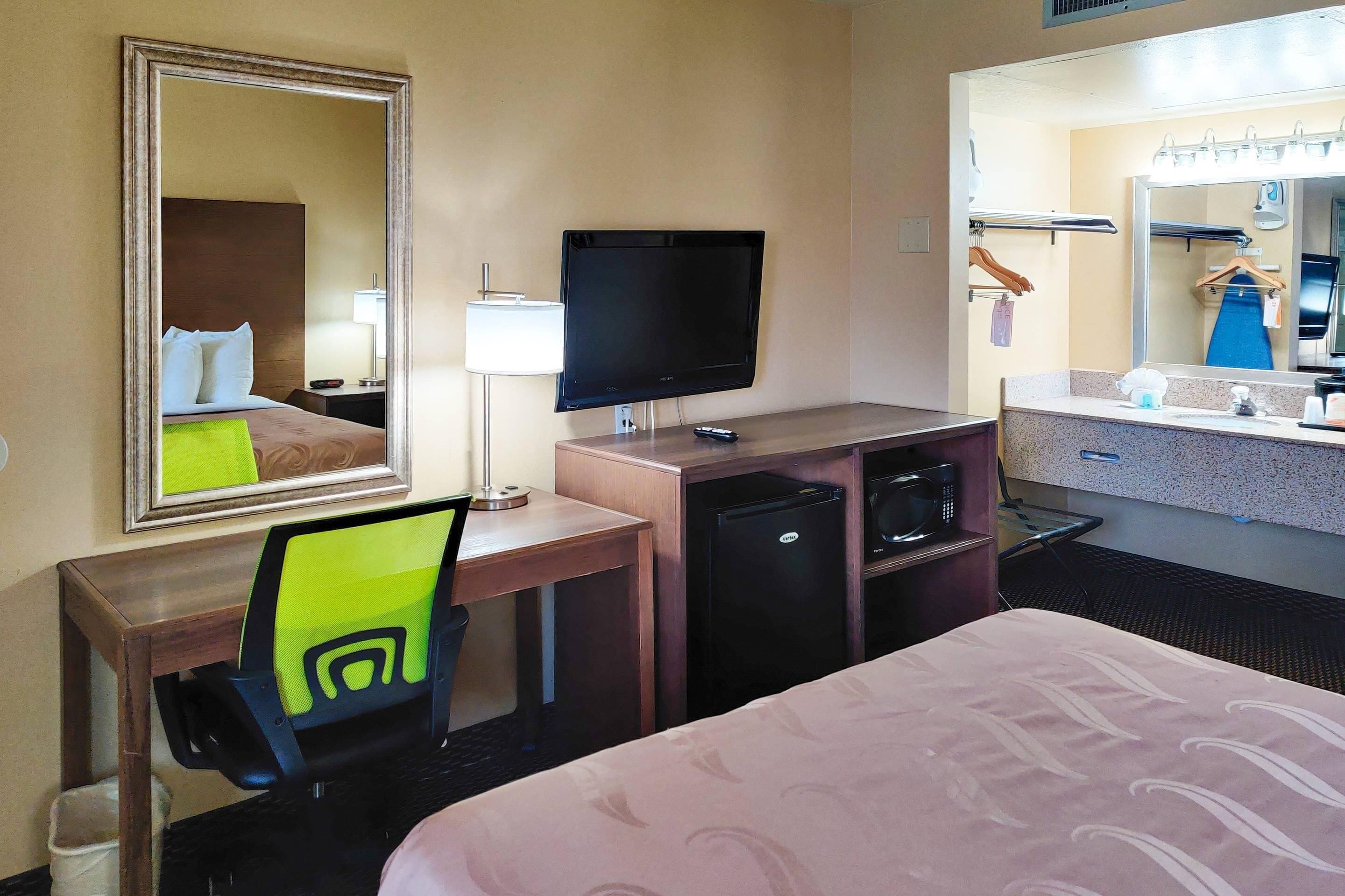 Een hotelkamer met een bed, een bureau met een computer, een televisie, een minibar en een badkamer met een wastafel en spiegel.