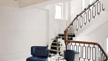 Een ruime kamer met een moderne trap, een blauwe fauteuil, een klein rond tafeltje en een vaas met witte bloemen.