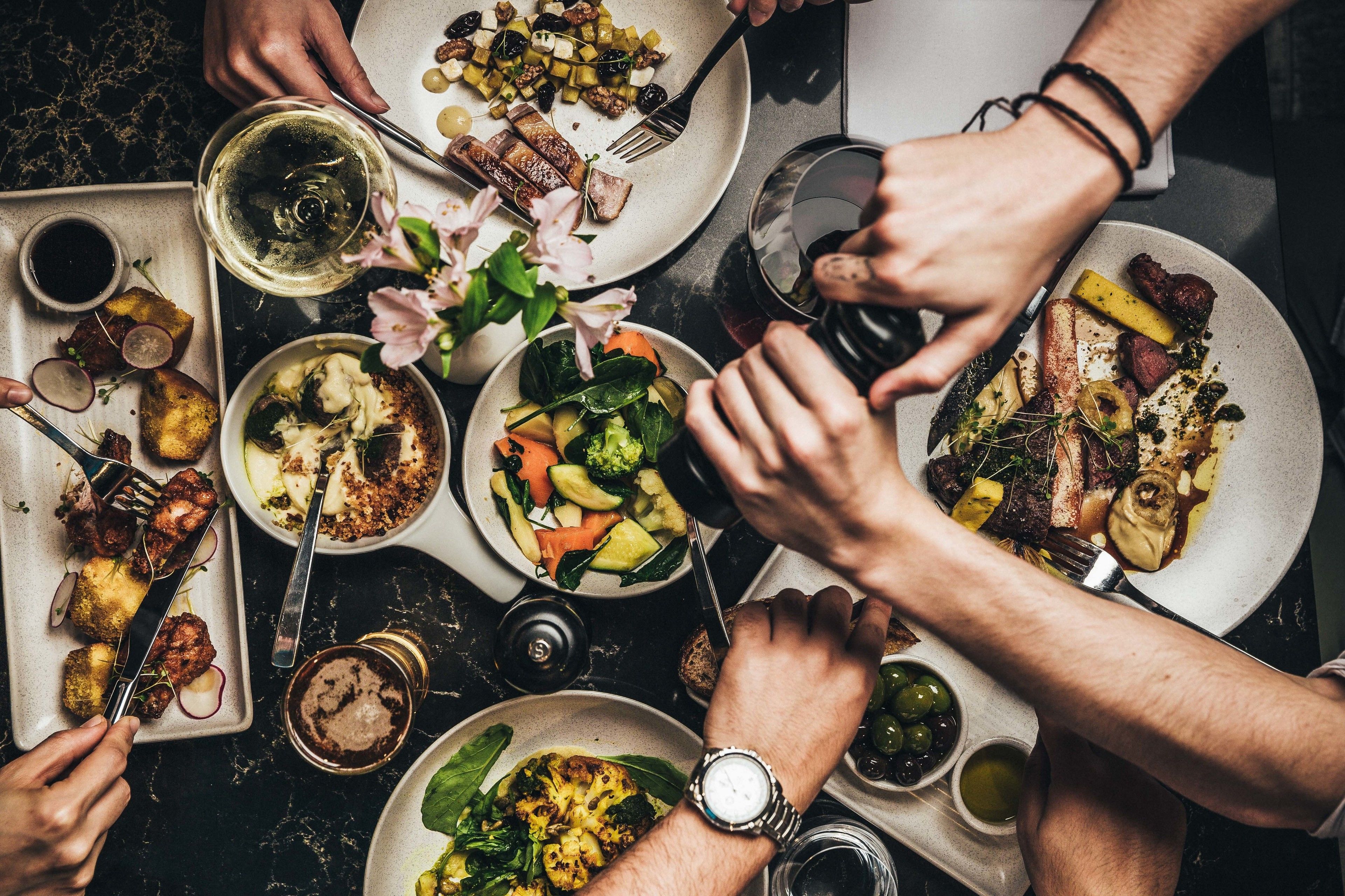 Um grupo de pessoas reunido em volta de uma mesa de jantar, com diversos pratos, bebidas e uma taça de vinho.