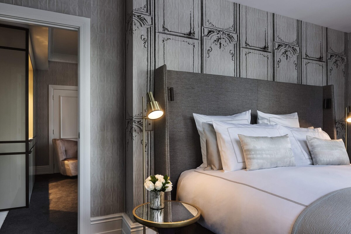 A hotel room with a large bed, a bedside table with a lamp, a chair, and a vase with flowers.