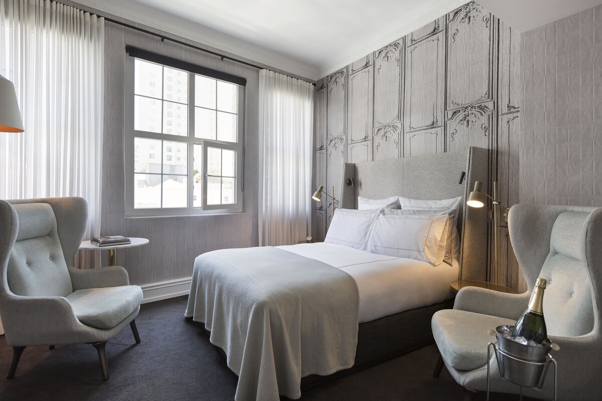 Une chambre d’hôtel avec un grand lit, deux fauteuils, une petite table et une fenêtre avec des rideaux.