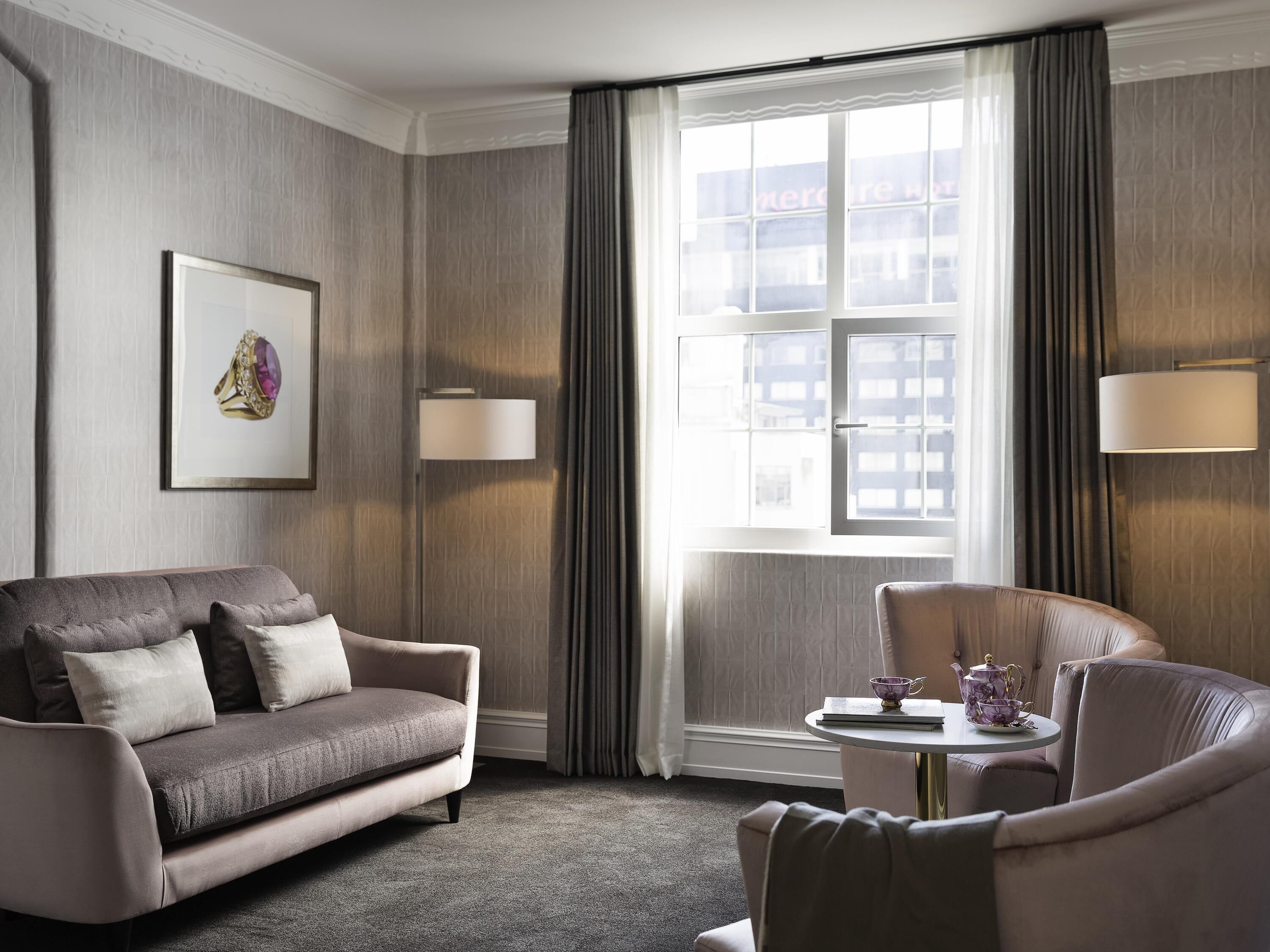 Een hotelkamer met een bank, fauteuil, een klein tafeltje met een theepot, en een raam met gordijnen.