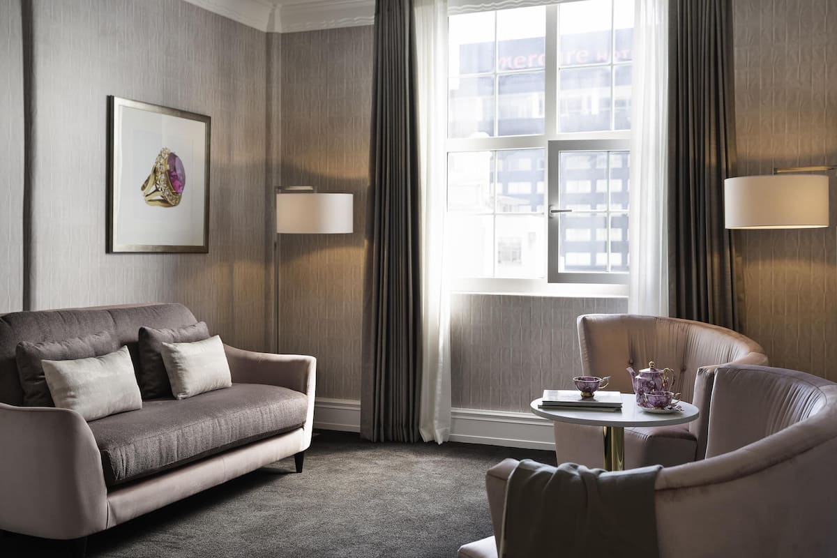 A hotel room with a sofa, armchair, a small table with a teapot, and a window with curtains.
