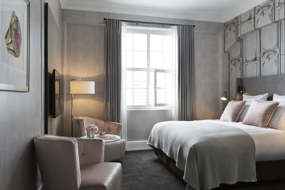 Une chambre d’hôtel avec un grand lit, un coin salon comprenant un fauteuil et une petite table, une fenêtre avec des rideaux et un téléviseur fixé au mur.