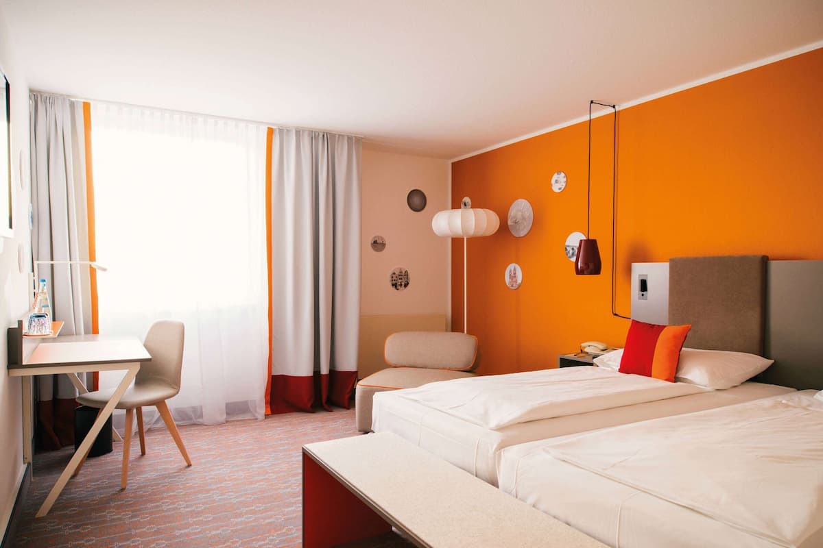 Une chambre d’hôtel avec deux lits, un bureau et une chaise. Il y a une fenêtre avec des rideaux, une horloge au mur et une table de chevet avec une lampe.