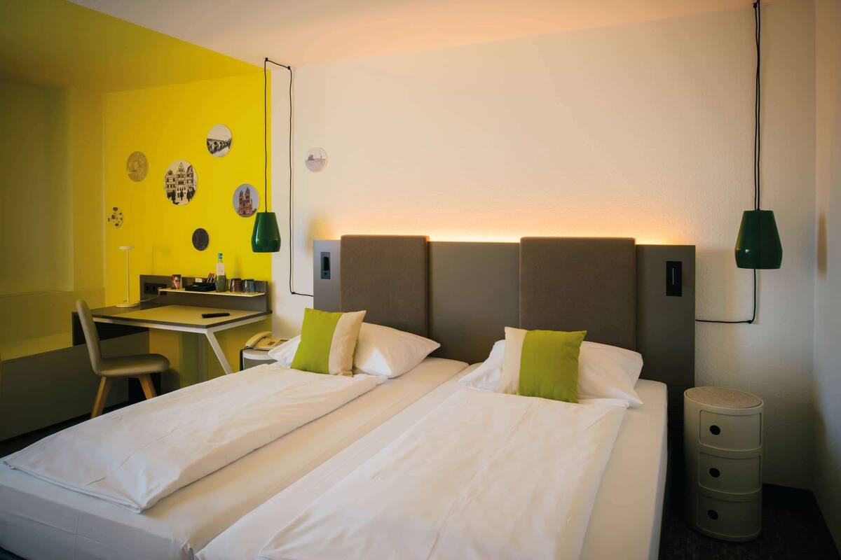 Une chambre d’hôtel avec deux lits, un bureau et une chaise. On y trouve des lampes fixées au mur et une petite table d’appoint.