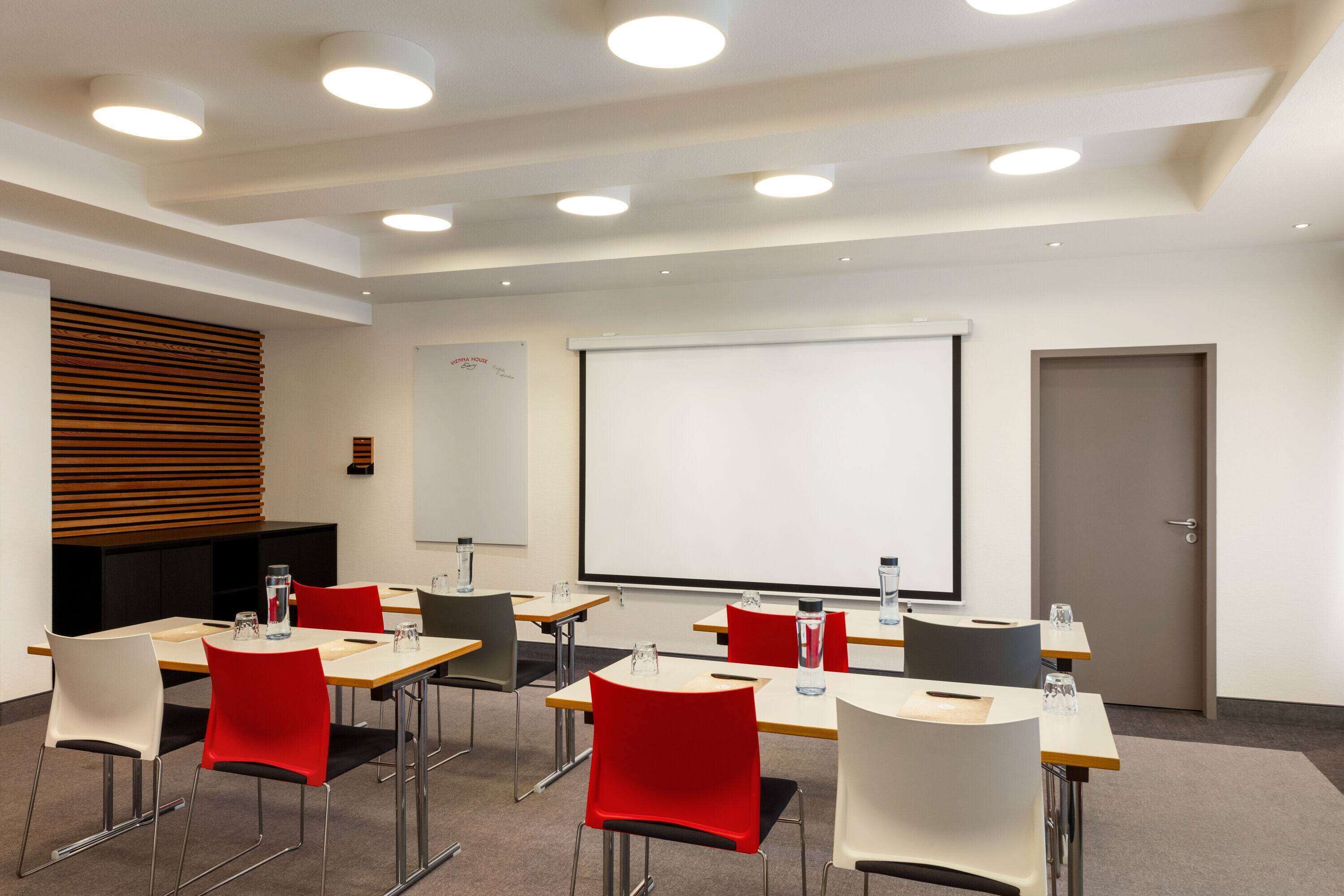 Un salón de conferencias con mesas, sillas, una pantalla de proyector y una pizarra.
