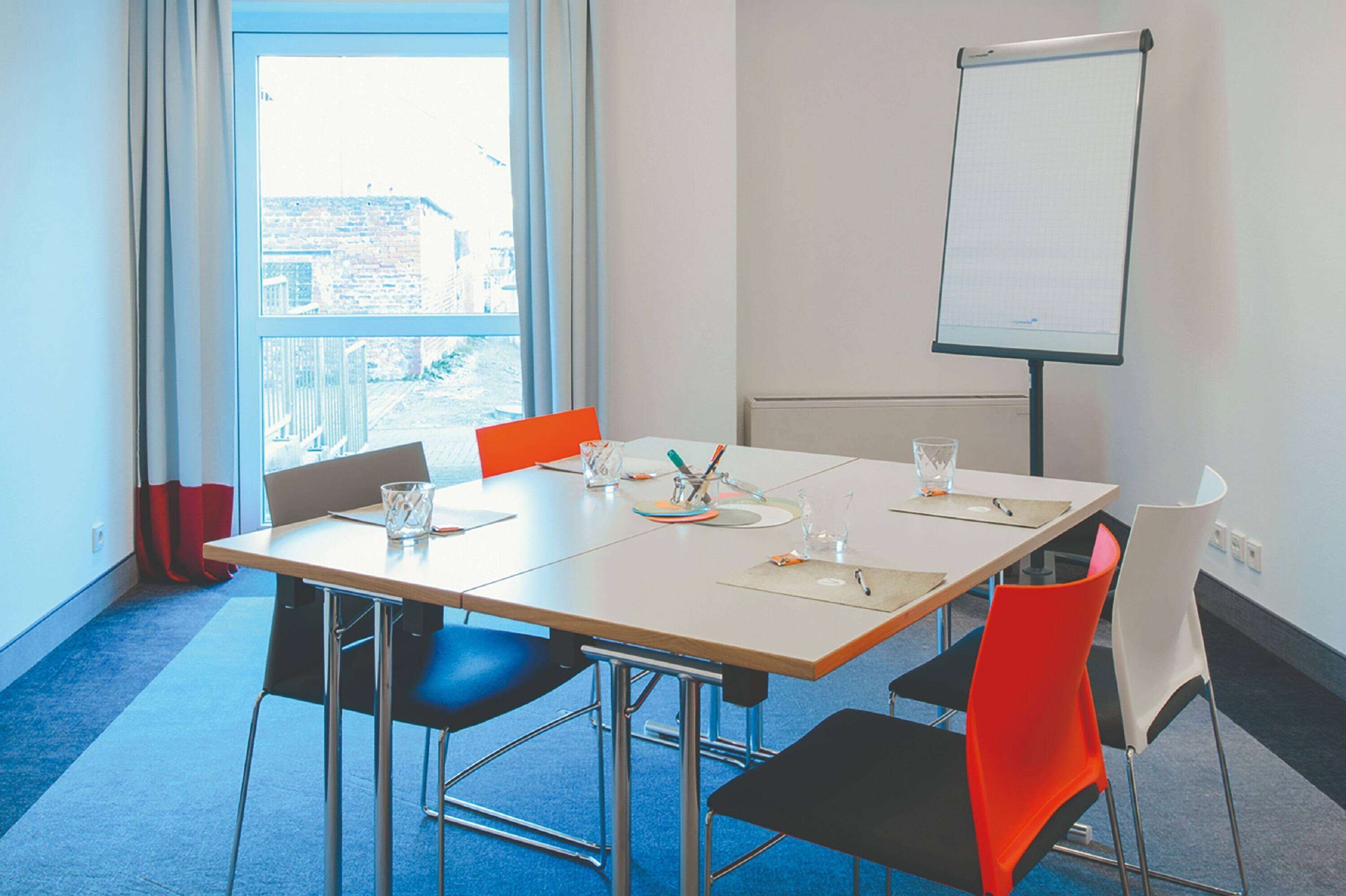 Un salón de conferencias con una gran mesa, cuatro sillas, una pizarra y vistas a edificios a través del ventanal.